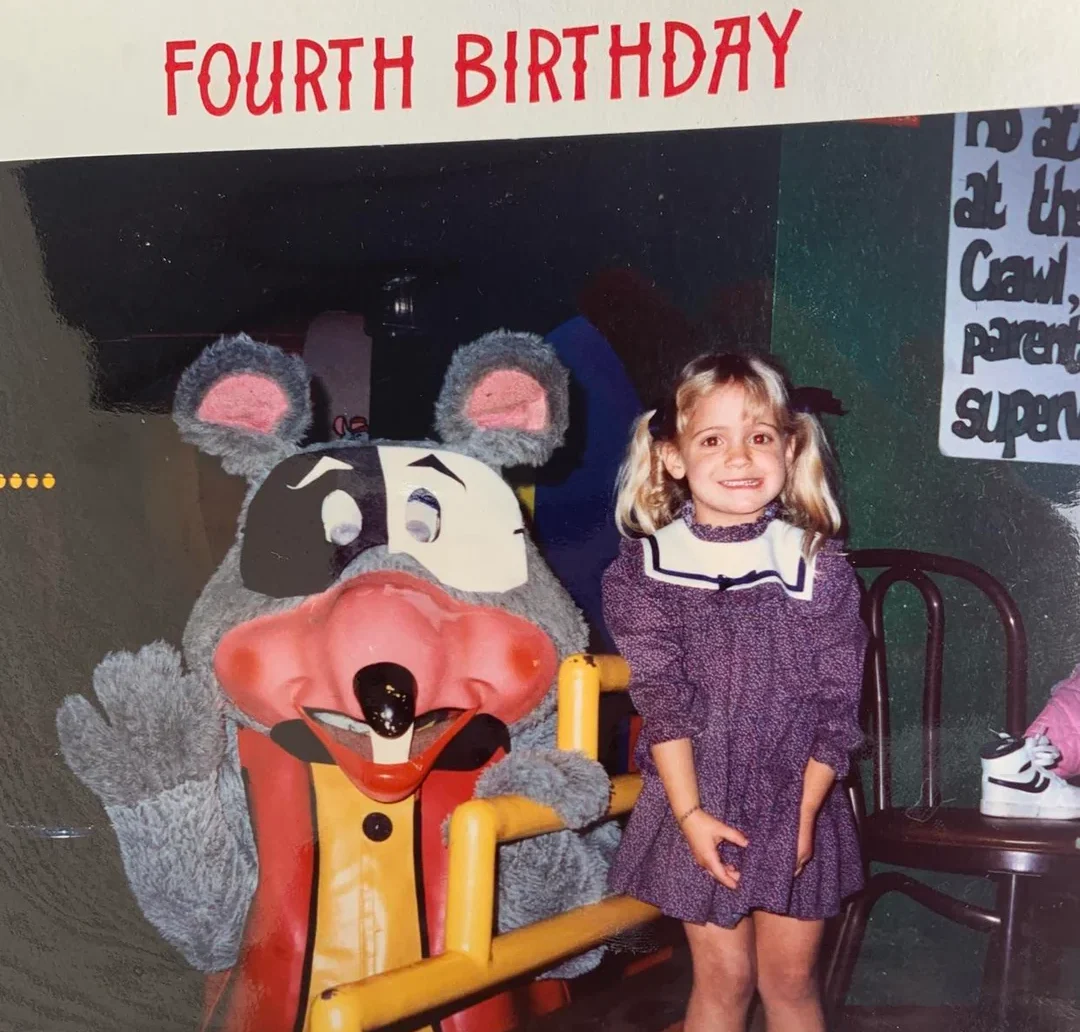 A young girl in a purple dress smiles next to a large Chuck E. Cheese mascot. Red “FOURTH BIRTHDAY” text appears above them. Colorful chairs and a painted sign are visible in the background.