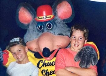 Two smiling children pose with the Chuck E. Cheese mascot, a large gray mouse in a yellow shirt and red cap, against a dark background. One child wears a white shirt and cap; the other wears a pink shirt.