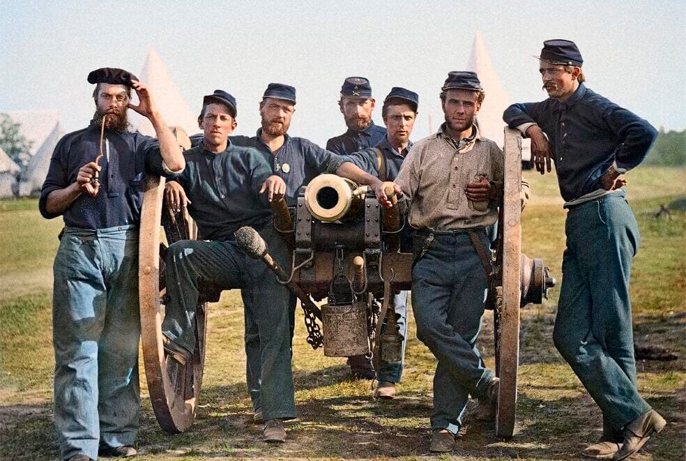 Six Union Civil War soldiers in blue uniforms pose casually around a cannon on grassy ground with white tents in the background, under a clear sky.