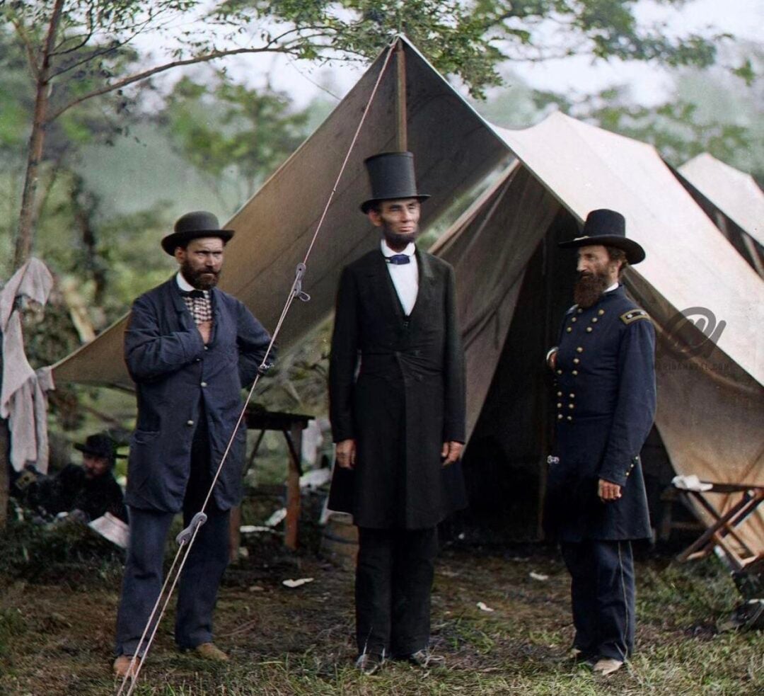 Three men stand in front of a canvas tent in a grassy, wooded area. The man in the center wears a tall stovepipe hat and black suit; the others wear 1800s attire, including a military uniform.