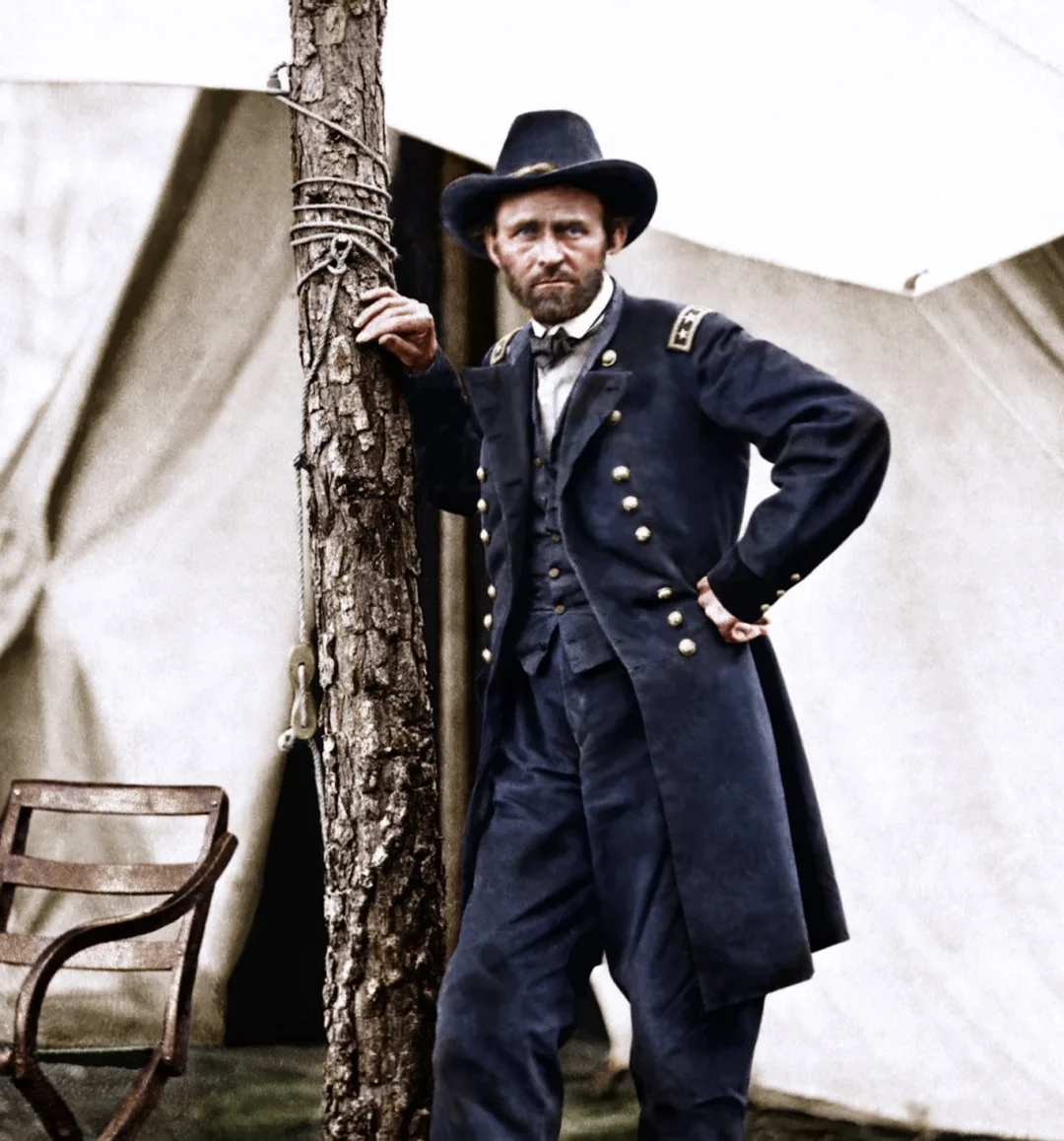 A colorized historical photo of a bearded man in a Civil War-era military uniform standing by a tree, with tents and a wooden chair in the background. Text at the bottom reads "Colorized By Victoria Heitshorn Studio Copyright 2021.