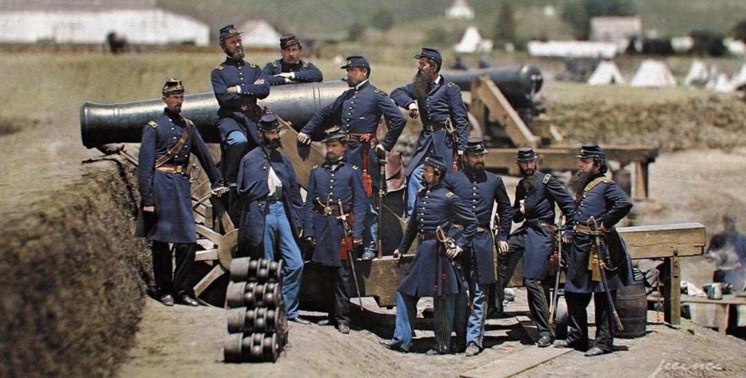 A group of Civil War-era soldiers in blue uniforms pose in front of a large cannon at a military camp, with tents and supplies visible in the background.