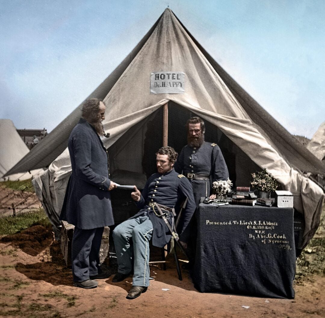Three men in Civil War-era military uniforms stand and sit outside a tent labeled "HOTEL." One man is seated, while the others stand beside a table with documents and a sign. The scene appears to be a camp.