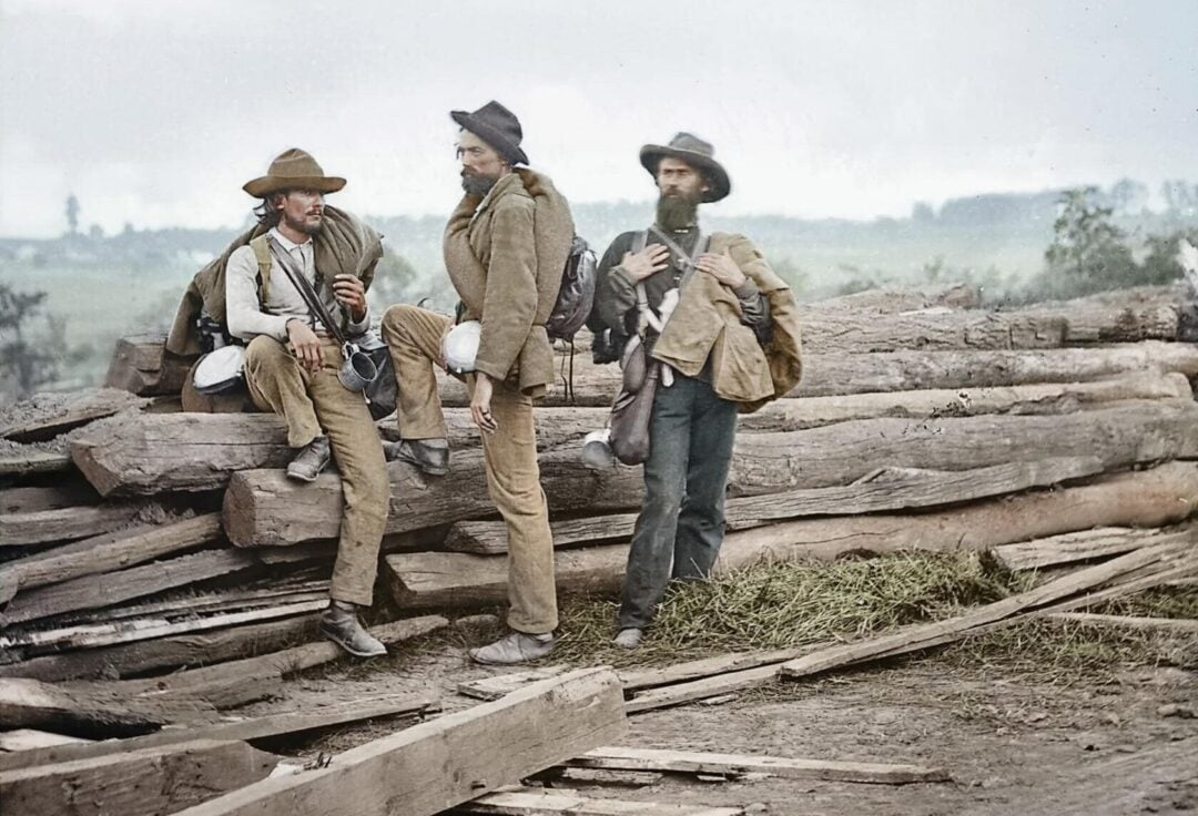 Three men in old-fashioned clothing, wearing hats and carrying bags, stand and sit on a pile of large wooden logs outdoors, with trees and a cloudy sky in the background.