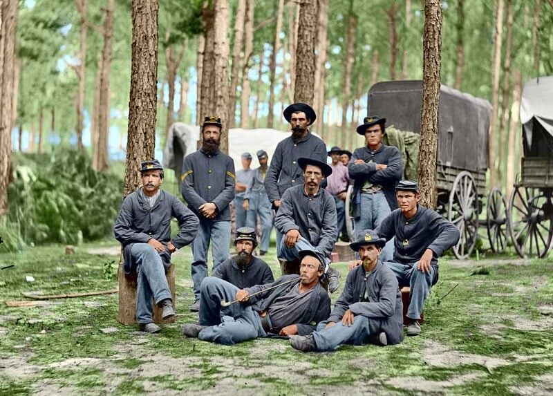 A group of Civil War soldiers in blue uniforms pose in a wooded area with covered wagons in the background; some are sitting or reclining on the ground, while others stand among the trees.