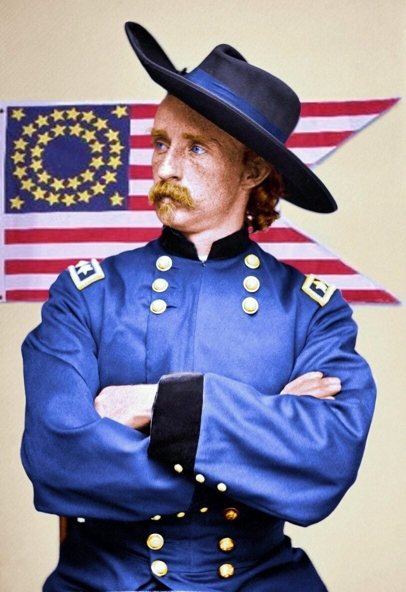 A man in a blue Civil War-era military uniform with gold buttons and shoulder insignia stands with arms crossed in front of a historic American flag. He has reddish hair, a mustache, and wears a wide-brimmed black hat.