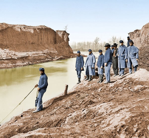 A group of men in blue Civil War-era uniforms stand on a muddy riverbank, while one man in front leans toward the water with a stick. The river runs between steep, bare earth embankments.