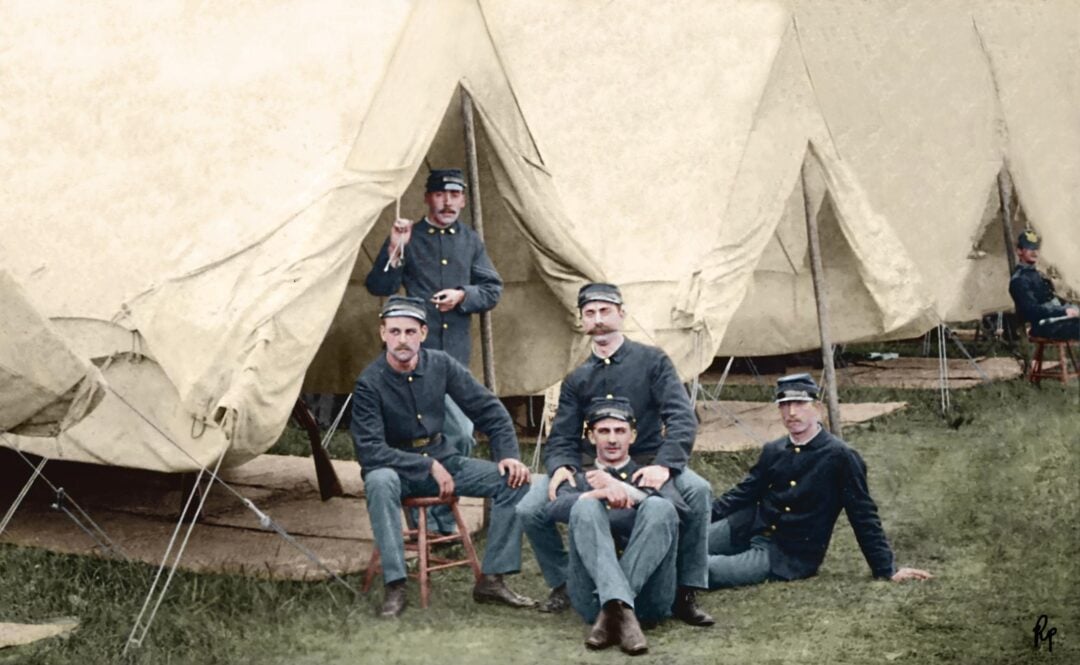 Five men in blue Civil War-era uniforms sit and pose in front of cream-colored canvas tents on grass, with three seated on chairs and two on the ground. Another uniformed man appears in the background near a tent.