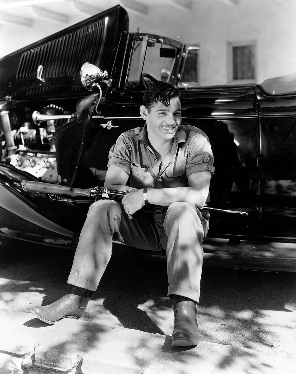 A man in a jumpsuit and boots sits on the running board of a vintage car, smiling and holding a rifle across his lap. The car’s hood is open and sunlight filters through the trees above.