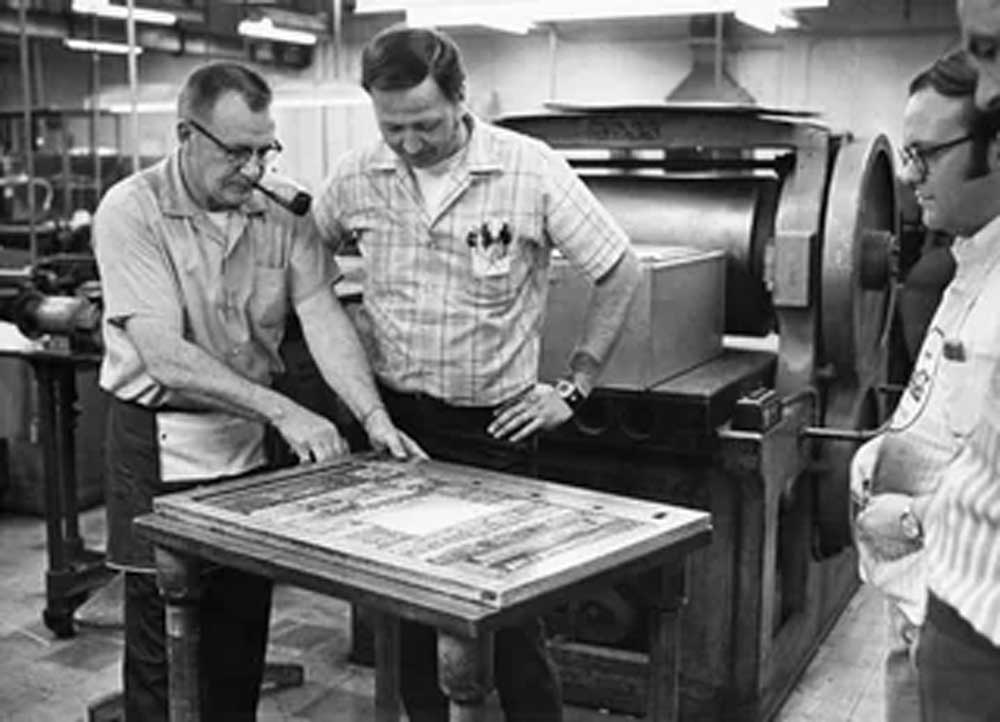 Three men stand around a table in a print shop, examining a layout. One man points at the table while others observe. Printing equipment and machinery are visible in the background.