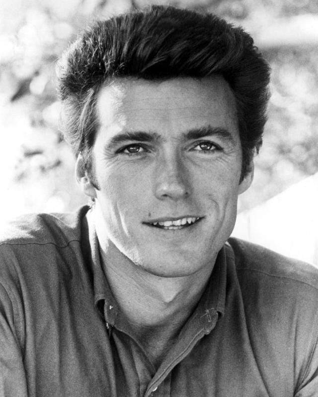 A black and white portrait of a man with dark, thick hair, wearing a button-up shirt, smiling slightly, and looking directly at the camera. The background is softly blurred with natural light.