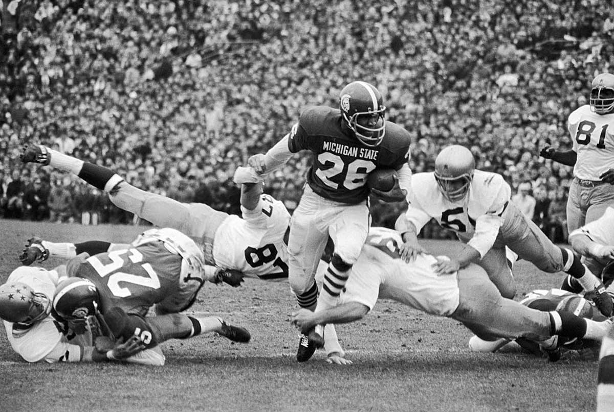 A Michigan State football player wearing number 26 runs with the ball as several opposing players dive and fall to the ground around him during a crowded game.