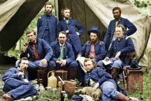 Nine men in blue Civil War-era military uniforms pose in front of a canvas tent. Some are seated or reclining, with mugs, bottles, and a basket on the ground, creating a relaxed camp scene.