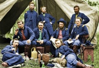 Nine men in blue Civil War-era military uniforms pose in front of a canvas tent. Some are seated or reclining, with mugs, bottles, and a basket on the ground, creating a relaxed camp scene.