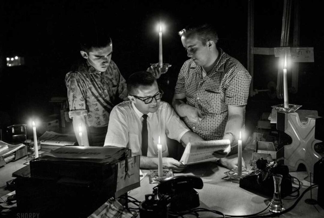 Three men work at a cluttered desk, reading and discussing papers by candlelight during a power outage; the scene is dimly lit, with candles providing the only source of light in the dark room.