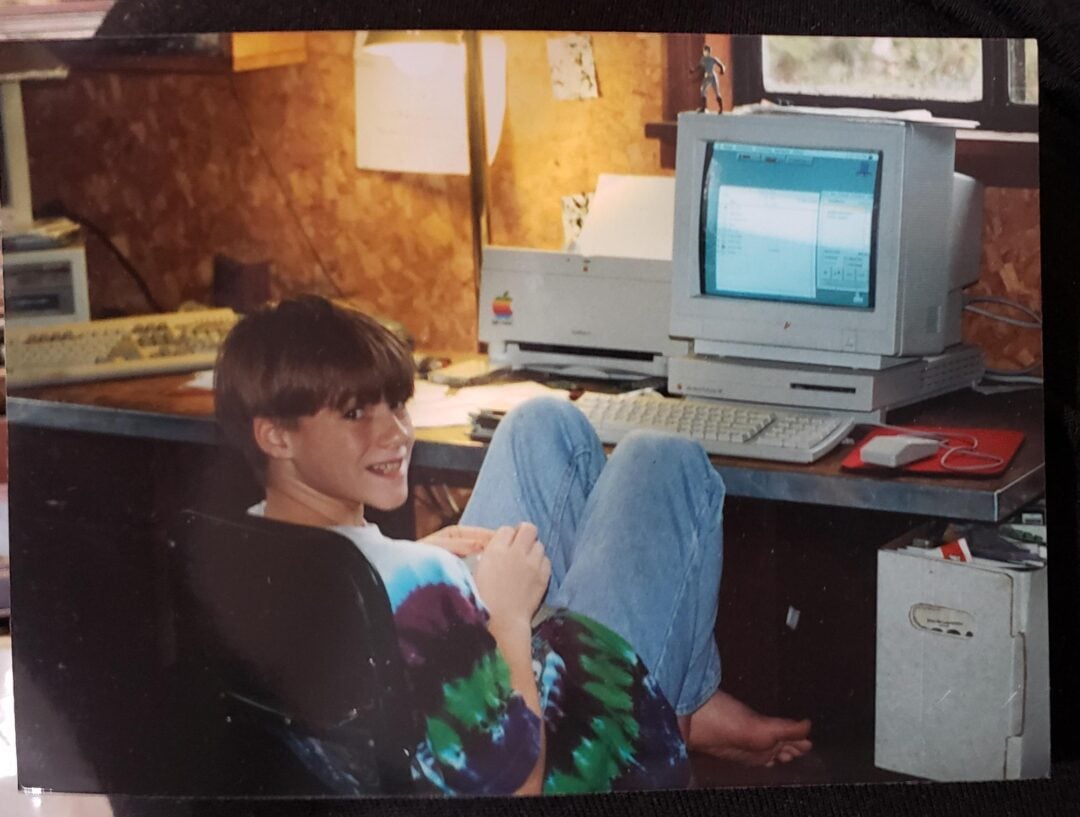 A boy in a tie-dye shirt sits barefoot at a desk with his legs up, smiling at the camera. An old Apple computer and retro keyboard are on the desk, with papers and office supplies nearby.