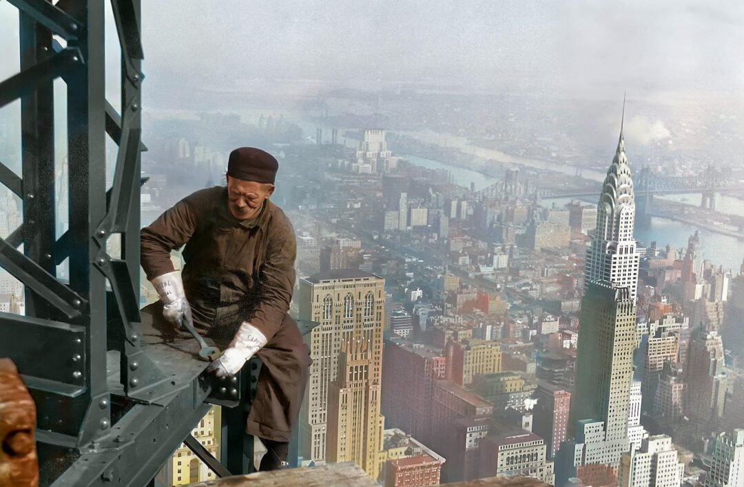 A construction worker in overalls and gloves secures steel beams high above New York City, with the Chrysler Building and East River visible in the background. The photo appears colorized and marked with @kenb.urns.