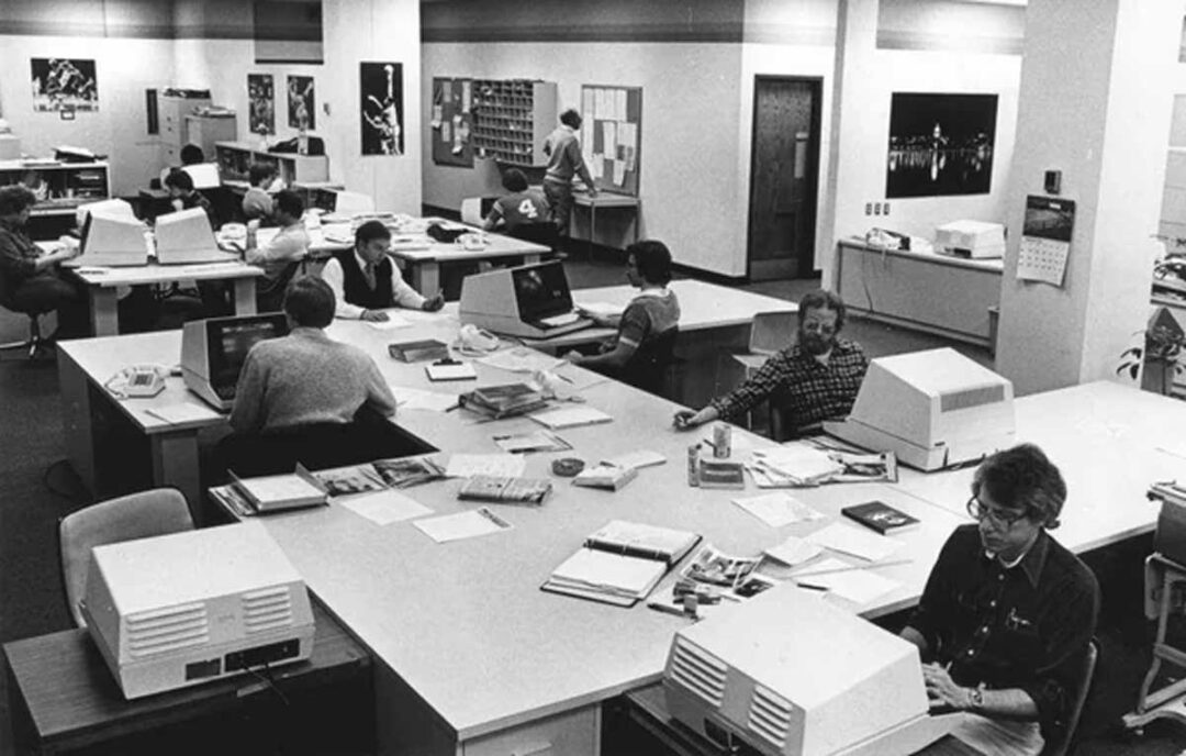 Black and white photo of a busy office in the 1980s, with people working at desks using early personal computers, surrounded by papers, books, and bulletin boards on the walls.