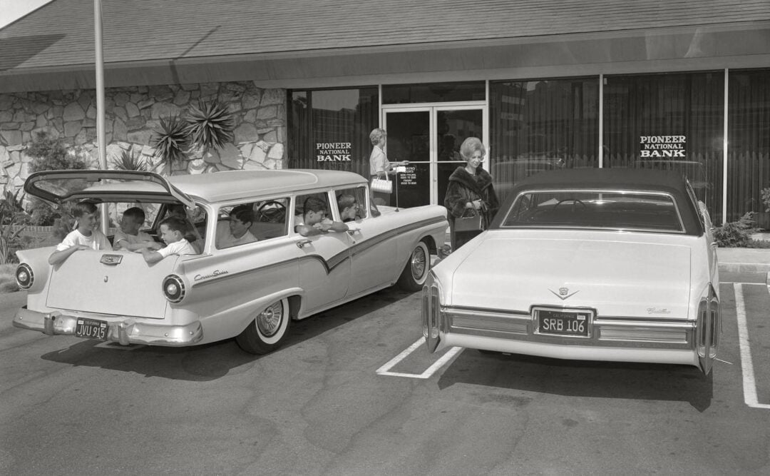 A black-and-white photo shows a woman standing beside a parked car outside "Pioneer National Bank" while five children sit in a nearby vintage station wagon, some looking out the windows.