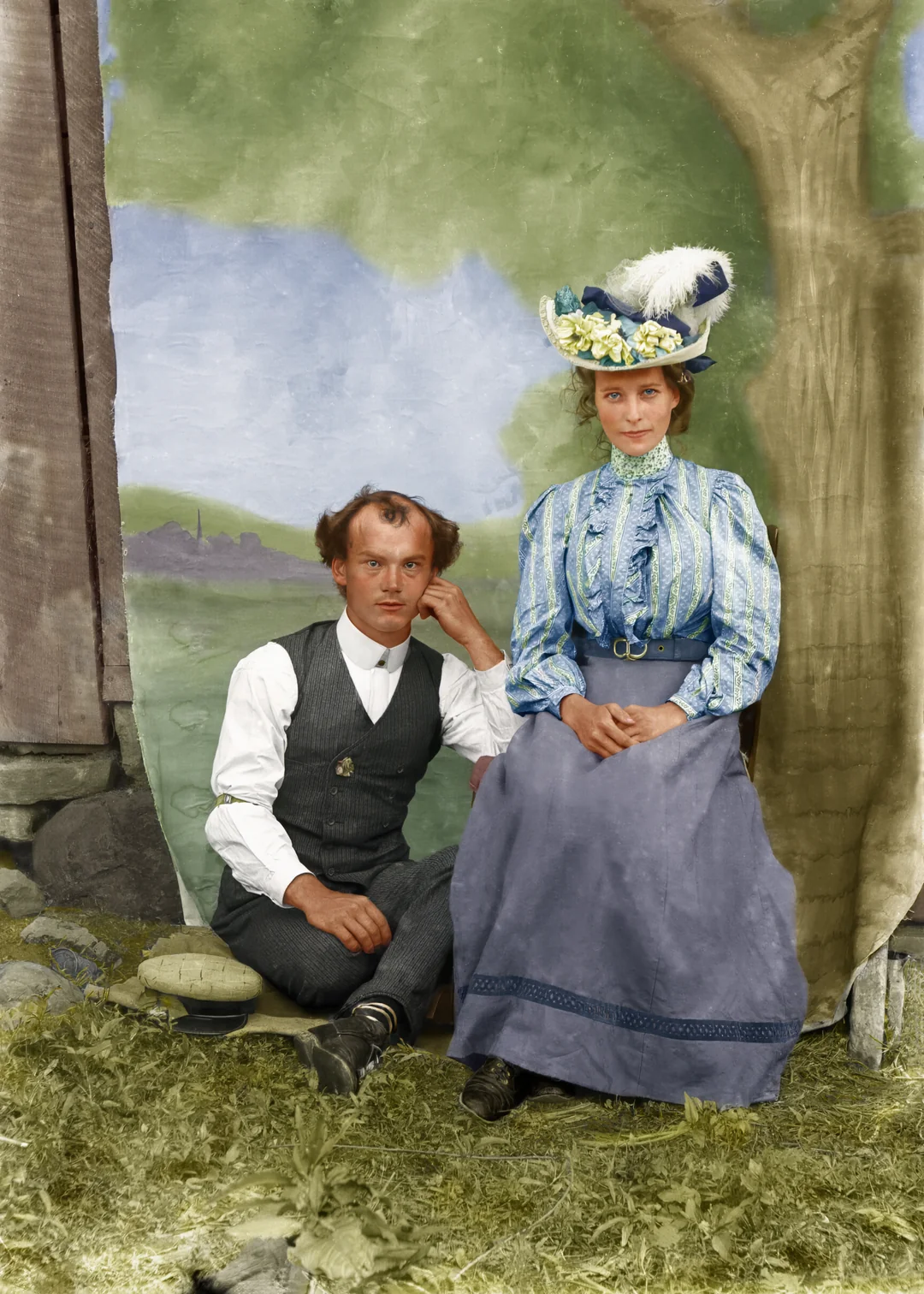 A man in a vest and tie sits on the grass beside a woman in a long blue skirt and a decorative hat, both posing in front of a painted outdoor backdrop and next to a tree.