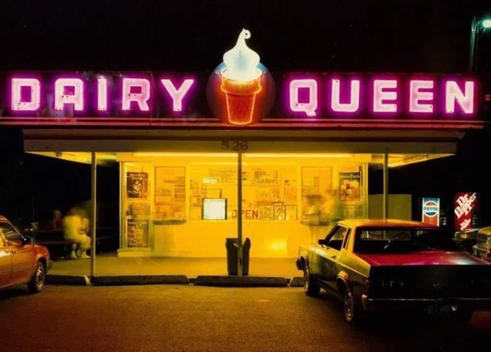 A brightly lit Dairy Queen restaurant at night, with a glowing neon sign and an ice cream cone logo. Two cars are parked in front, and the inside is illuminated with people visible through the windows.