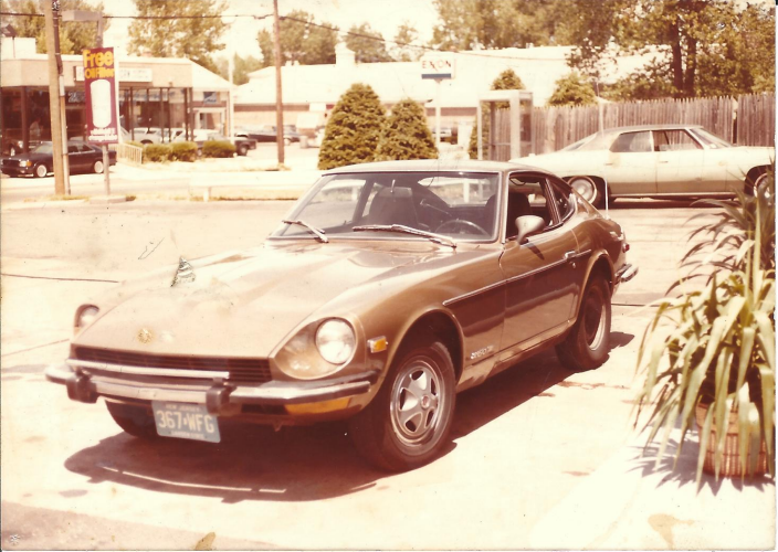 A brown vintage Datsun 240Z sports car is parked in a lot on a sunny day, with plants in the foreground and shops, another car, and trees visible in the background.