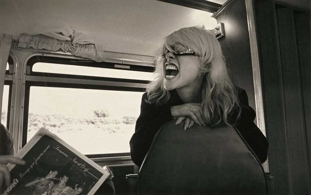 A woman with light hair and sunglasses leans over the back of a bus seat, mouth wide open as if shouting or laughing. Sunlight streams through the window behind her. Another person holds a magazine in the foreground.