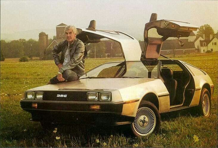 A man sits on the hood of a stainless steel DeLorean car with its gull-wing doors open, parked in a grassy field with buildings and trees in the background.