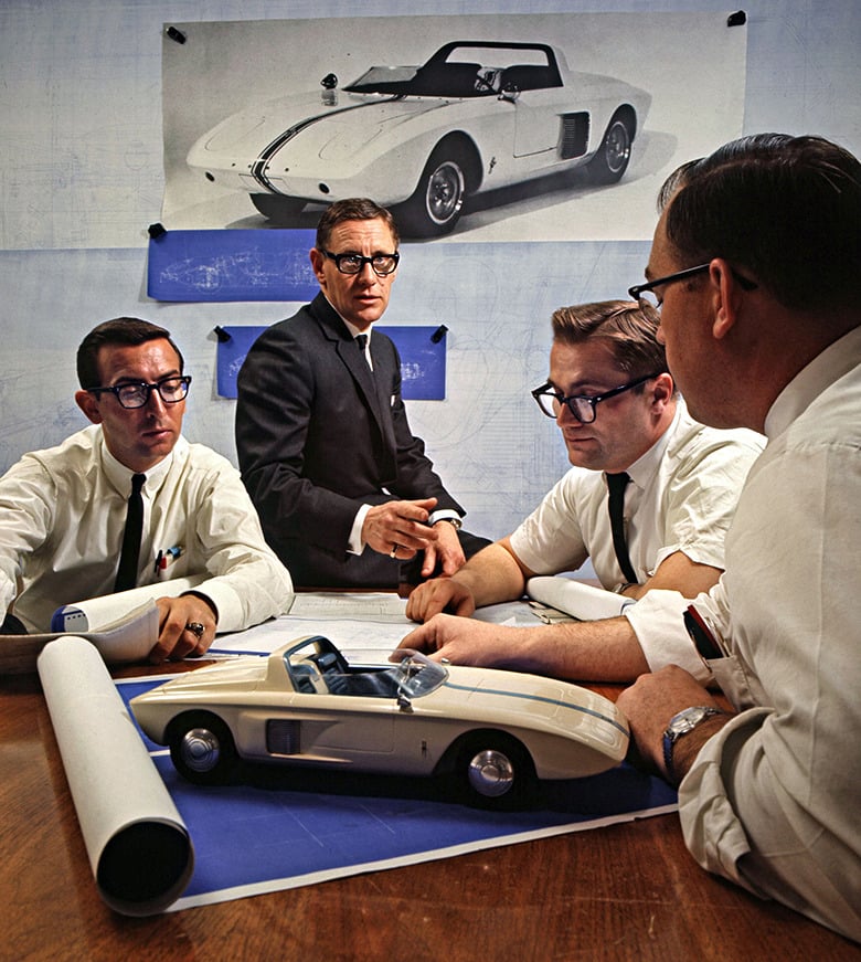 Four men in white shirts and black glasses sit at a table with blueprints and a model car, discussing a car design. A large drawing of the same car is pinned to the wall behind them.