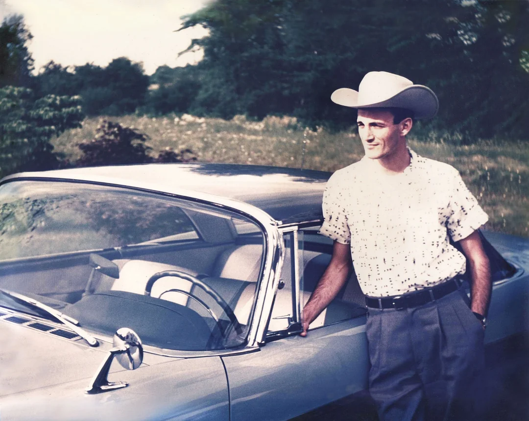 A man wearing a cowboy hat and a patterned short-sleeve shirt stands by the open door of a classic car in a grassy outdoor setting, with trees and foliage in the background.