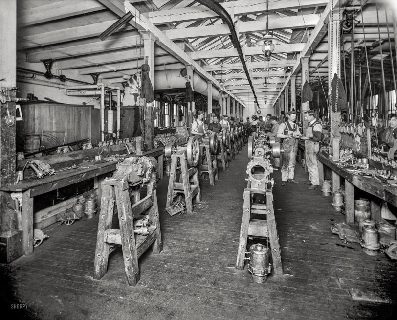Historic black-and-white photo of workers assembling machinery in a large, crowded factory with wooden floors, beams, and workbenches lined with tools and metal parts. Workers wear uniforms, focused on their tasks.