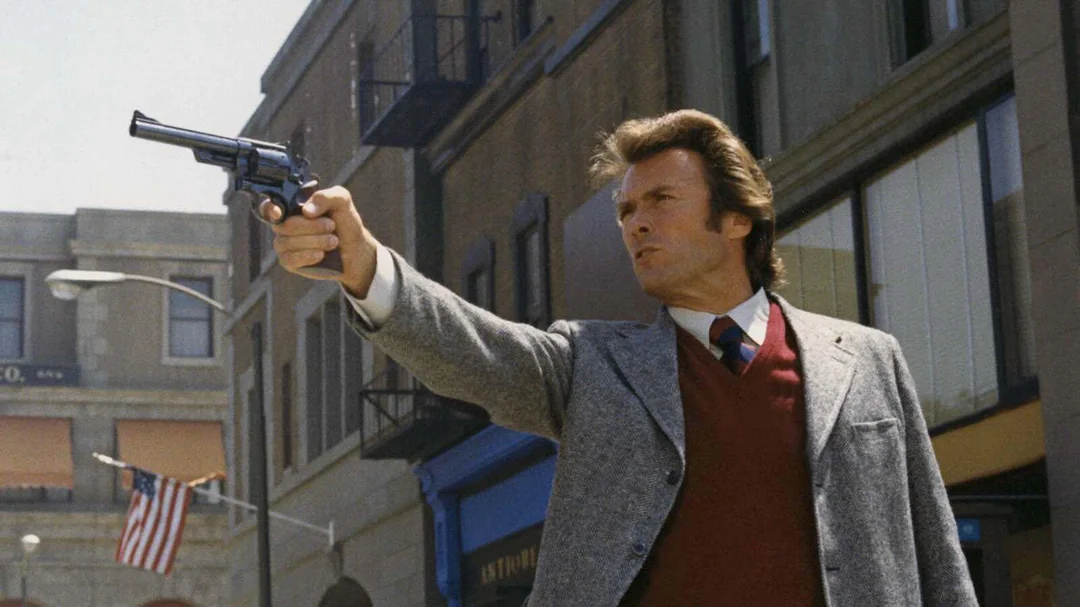 A man in a suit and red sweater aims a revolver on a city street, with buildings and an American flag visible in the background.