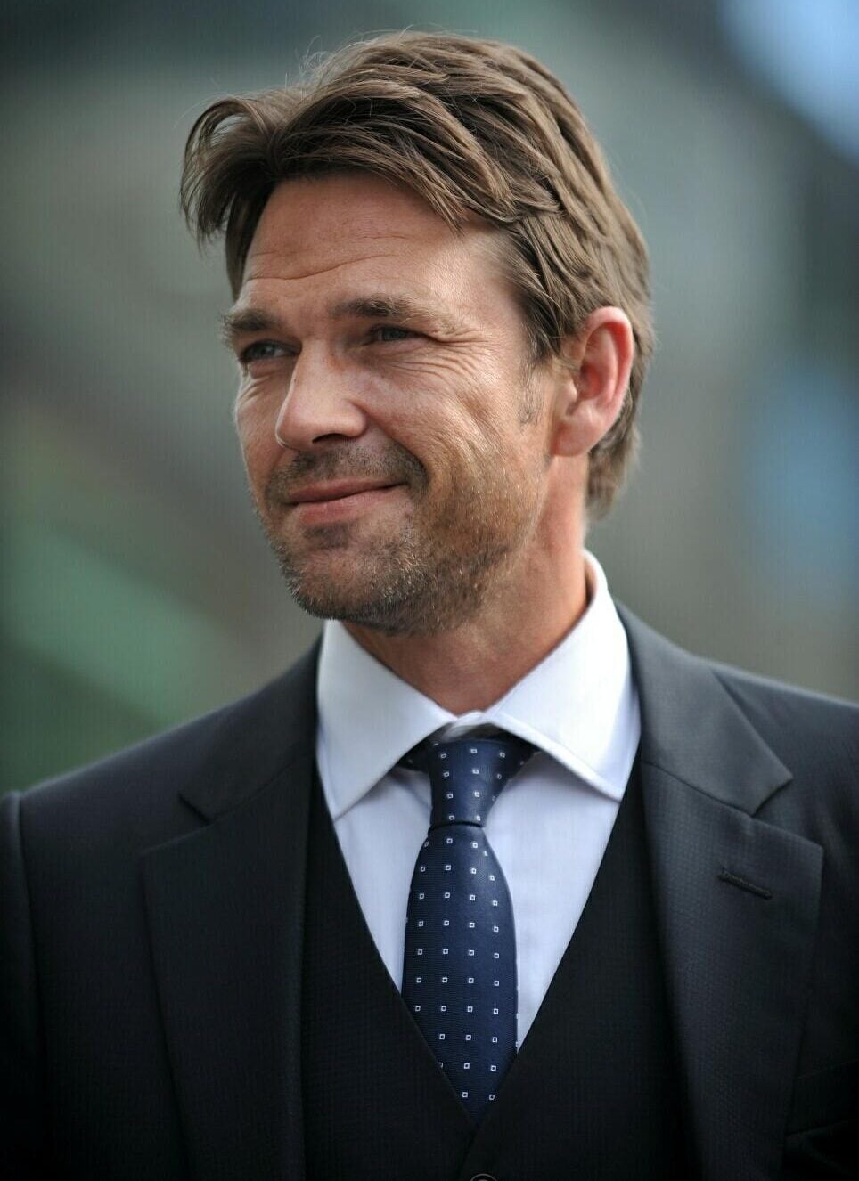 A man with short brown hair and light stubble is wearing a dark suit, a white shirt, and a navy blue tie with small white dots. He is smiling softly and standing outdoors with a blurred background.