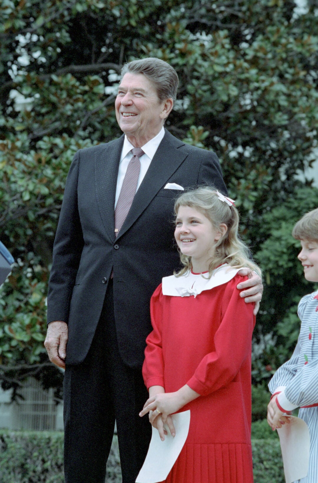 A man in a dark suit stands outdoors with his arm around a smiling young girl in a red dress. Another child stands nearby. Trees and greenery are visible in the background.