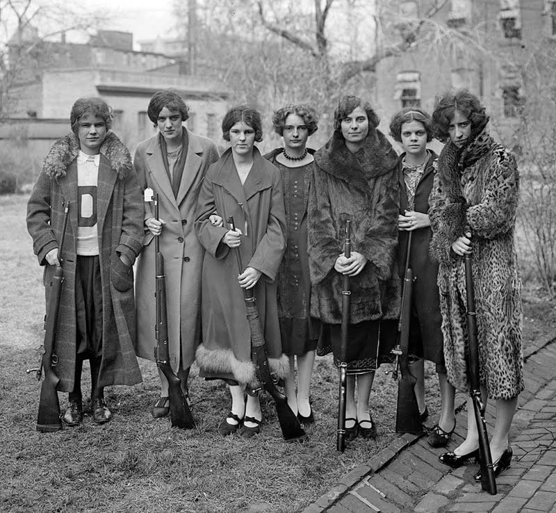 Seven women in 1920s-style clothing stand outdoors, holding rifles. The group is posed in a line, with serious expressions, in a yard with bare trees and buildings in the background.