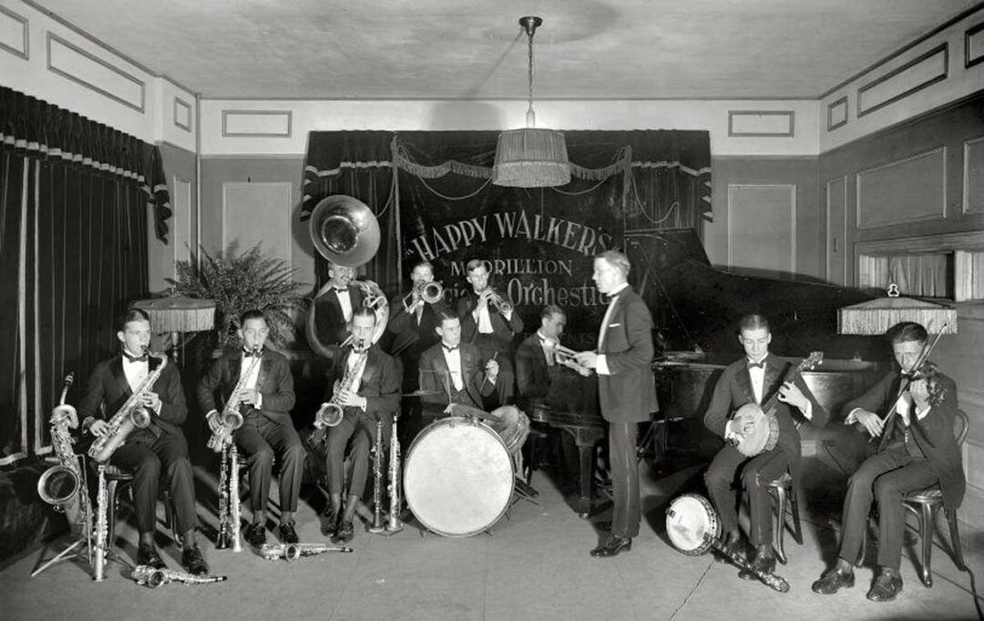 A black-and-white photo of a jazz band in tuxedos playing various instruments, including saxophones, trumpet, drums, piano, and banjo, on a stage with a banner reading "Happy Walker’s Pavilion Original Orchestra.