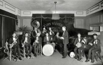 A black-and-white photo of a jazz band in tuxedos playing various instruments, including saxophones, trumpet, drums, piano, and banjo, on a stage with a banner reading "Happy Walker&rsquo;s Pavilion Original Orchestra.
