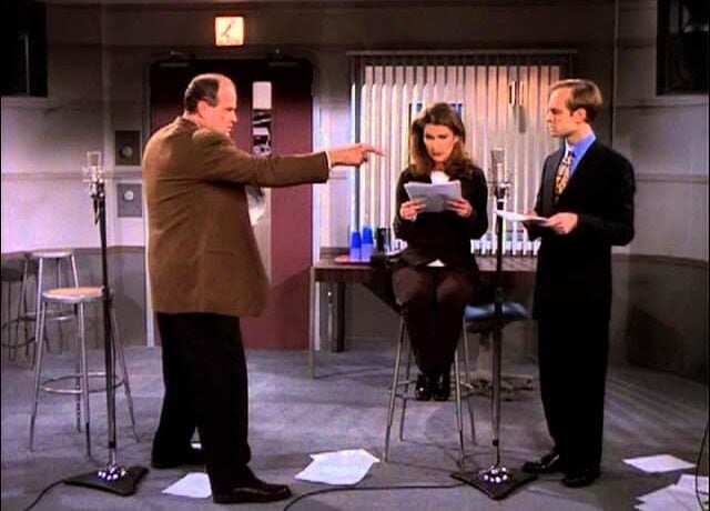 Three people are in a recording studio. A man stands and points while another man and a woman, both holding scripts, stand and sit near microphones. Papers are scattered on the floor.