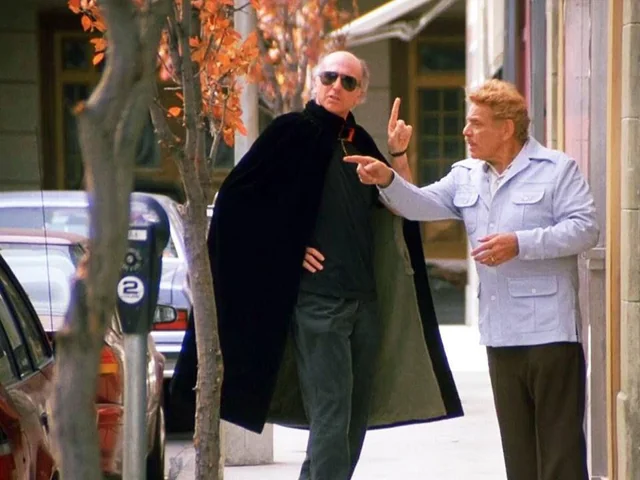 Two men stand on a city sidewalk; one wears sunglasses and a dark cape, the other gestures with his hand while speaking. Autumn leaves hang on nearby trees and parked cars line the street.