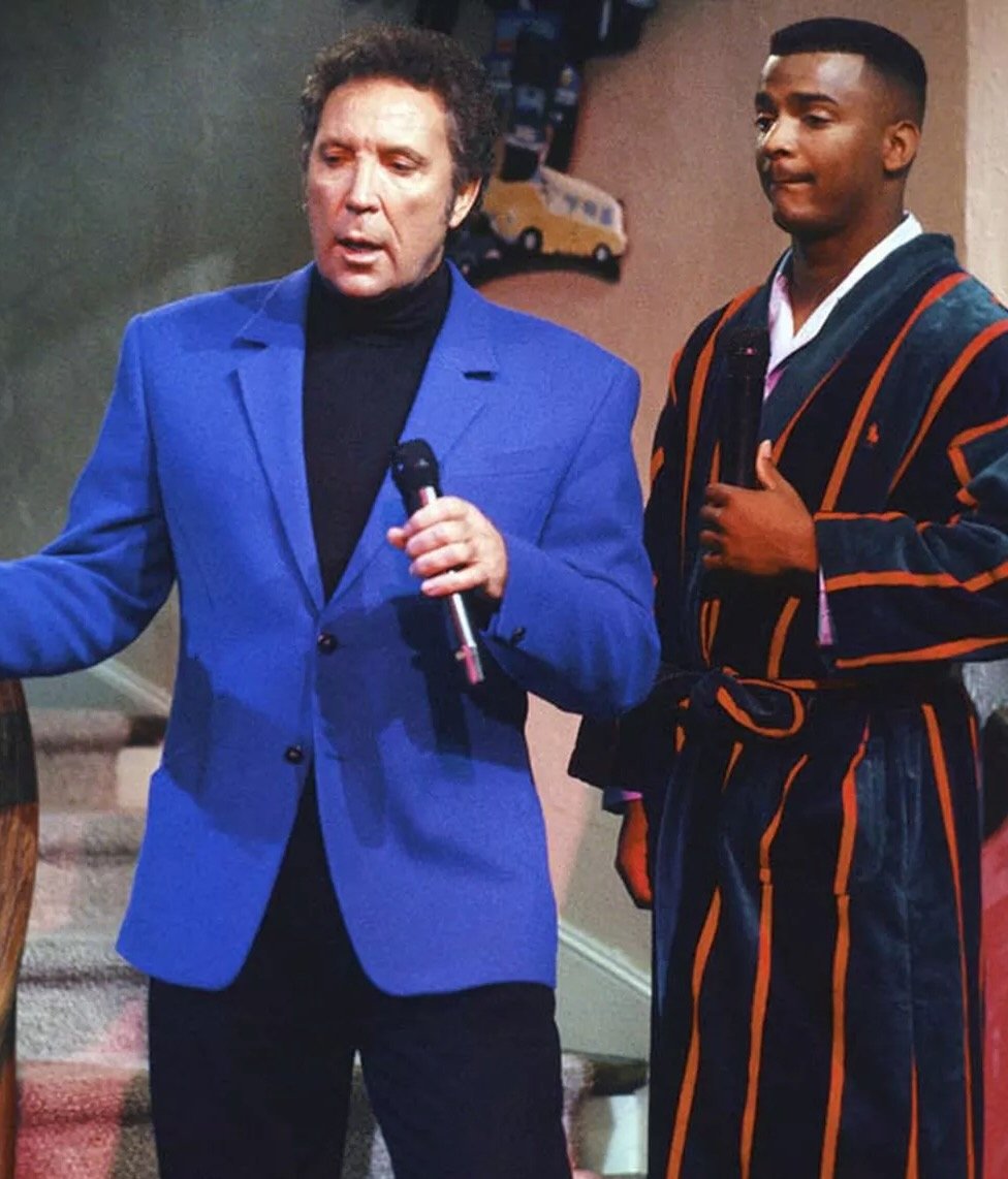 A man in a bright blue blazer holds a microphone while standing next to a young man in a striped robe, both indoors near a staircase with toy vehicles on the wall behind them.