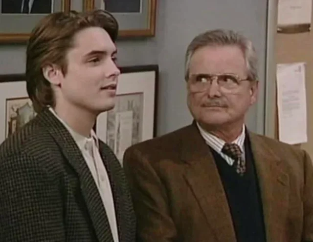 A young man with medium-length brown hair stands next to an older man with gray hair, glasses, and a mustache. Both are wearing jackets and ties, and they appear to be indoors in an office or classroom setting.