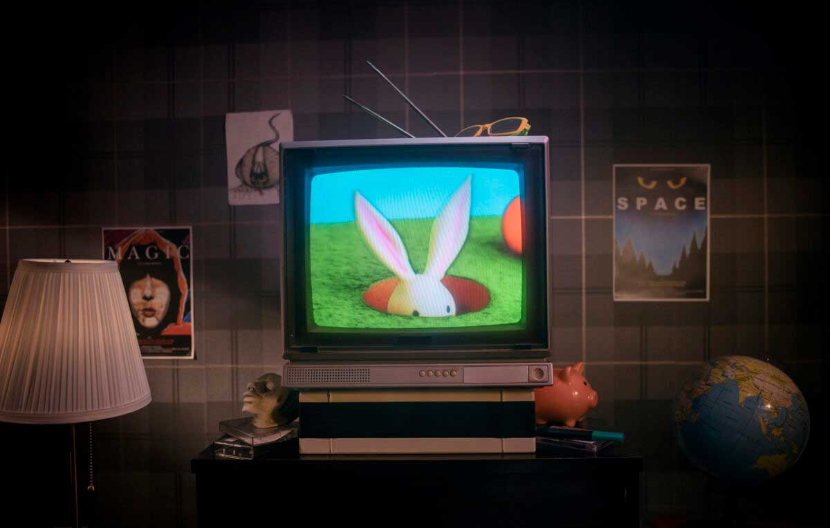A vintage TV displays a cartoon rabbit emerging from a hole. The TV sits on a table with a lamp, globe, piggy bank, and skull nearby. Posters and decorations are on the plaid-patterned wall behind.
