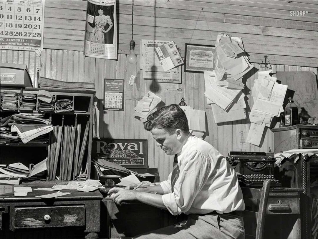 A man sits at a cluttered desk in an office, surrounded by scattered papers, files, and calendars pinned on the wooden wall behind him. He appears focused as he writes or examines documents.