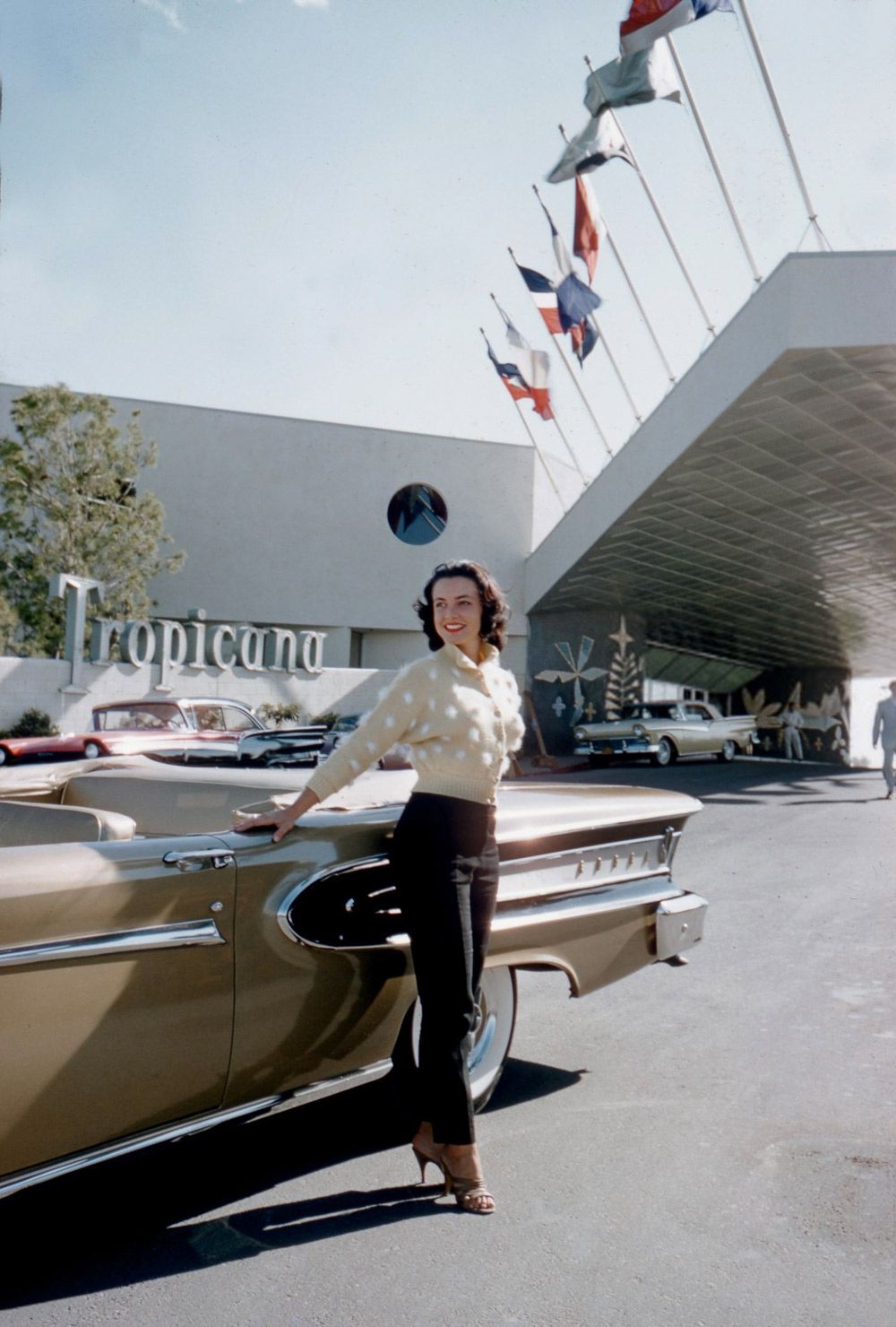 A woman in a white sweater and black pants poses beside a classic gold car in front of the Tropicana hotel, with flags and vintage cars in the background on a sunny day.