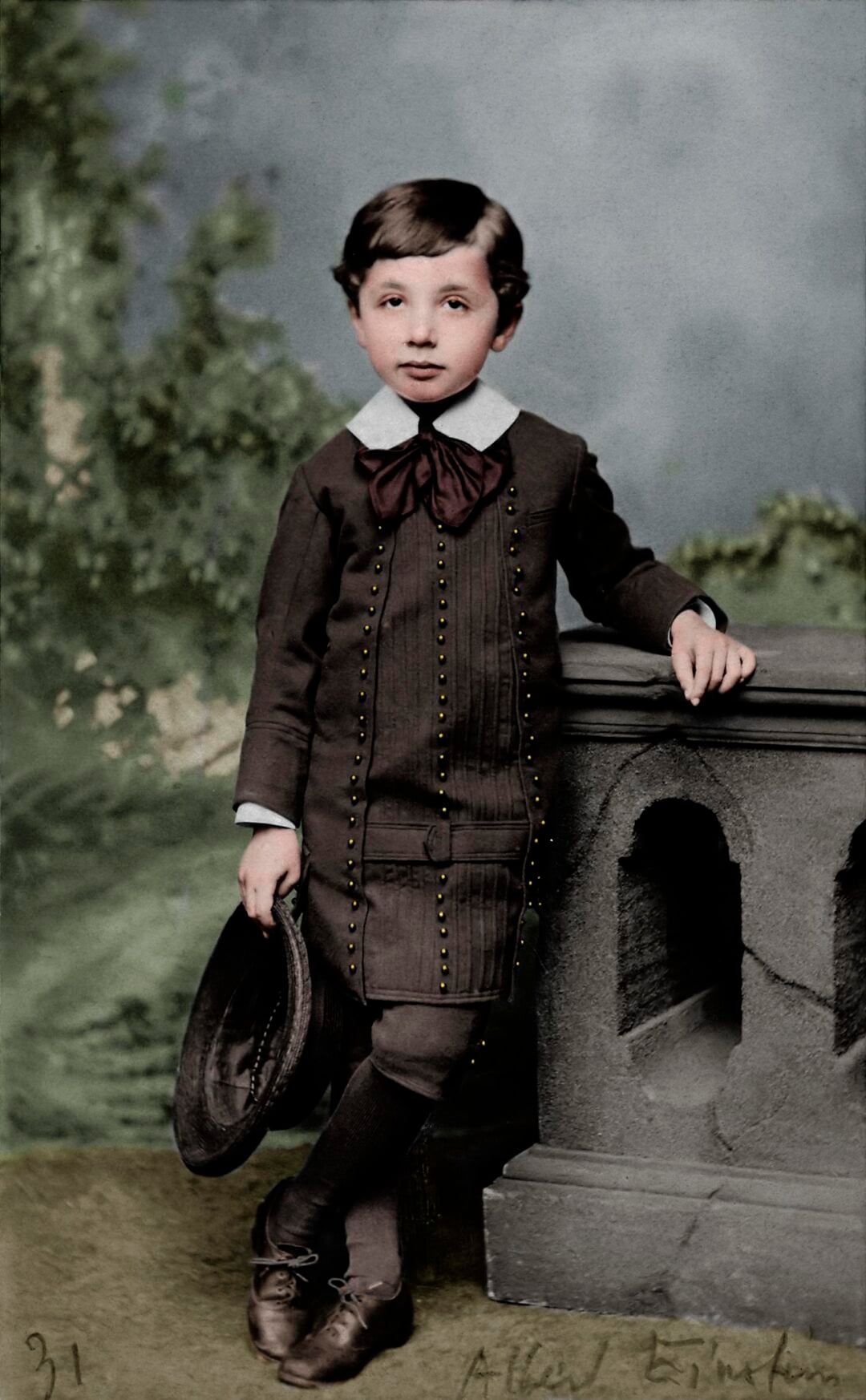 A young boy with dark hair, dressed in a brown 19th-century suit with a large bow and holding a hat, stands by a stone railing with a nature backdrop.