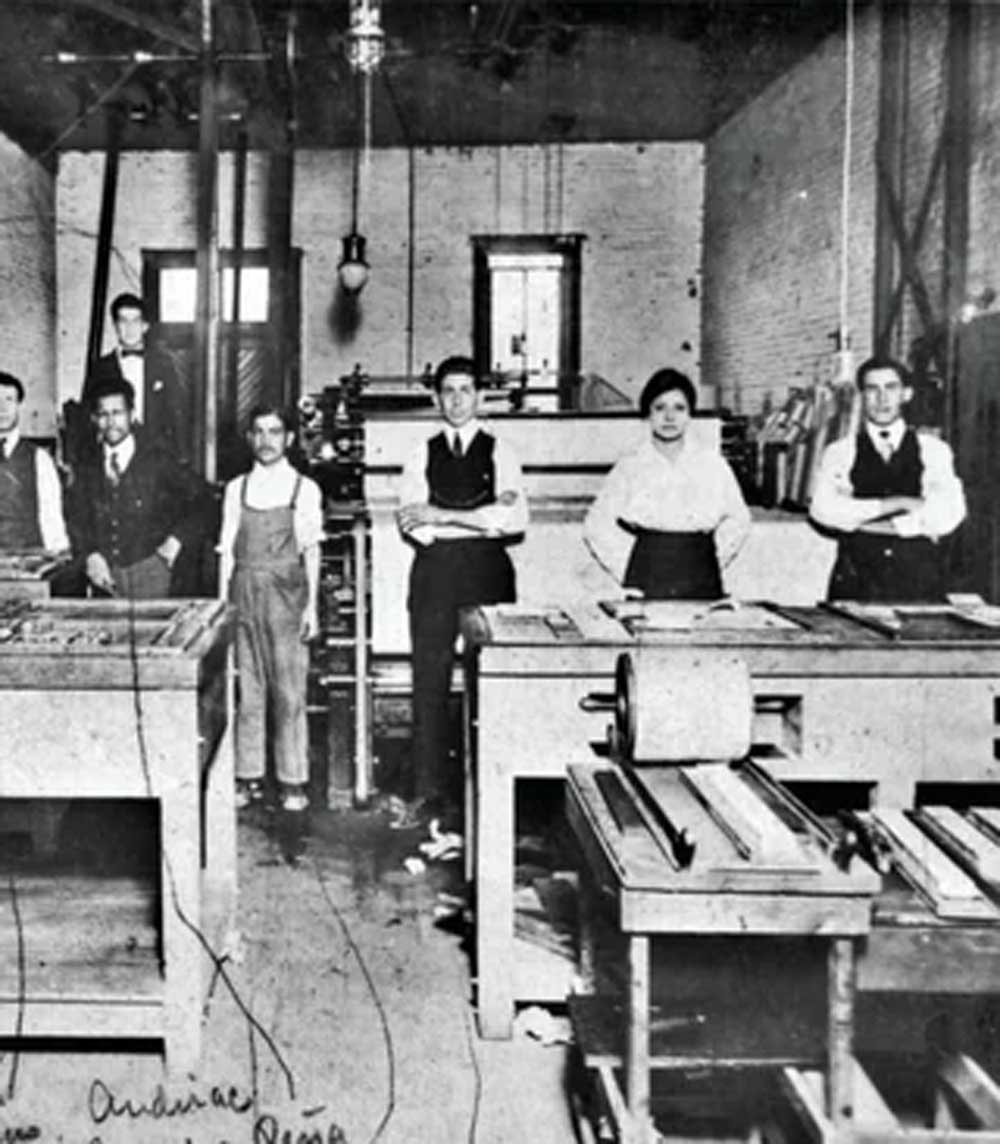 A black and white photo of men and one woman standing in a print shop with large printing presses and workbenches, wearing early 20th-century work attire.