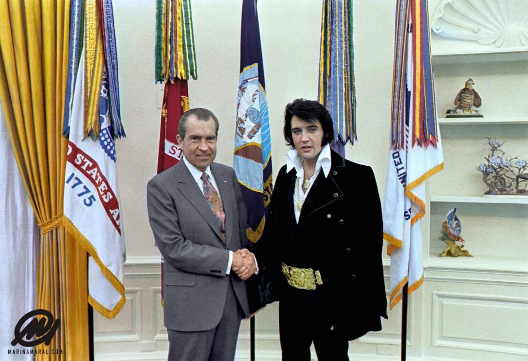 Richard Nixon and Elvis Presley shake hands in front of several U.S. military flags in a formal, official setting. Both are dressed formally; Nixon in a suit, Presley in a black outfit with a large belt.