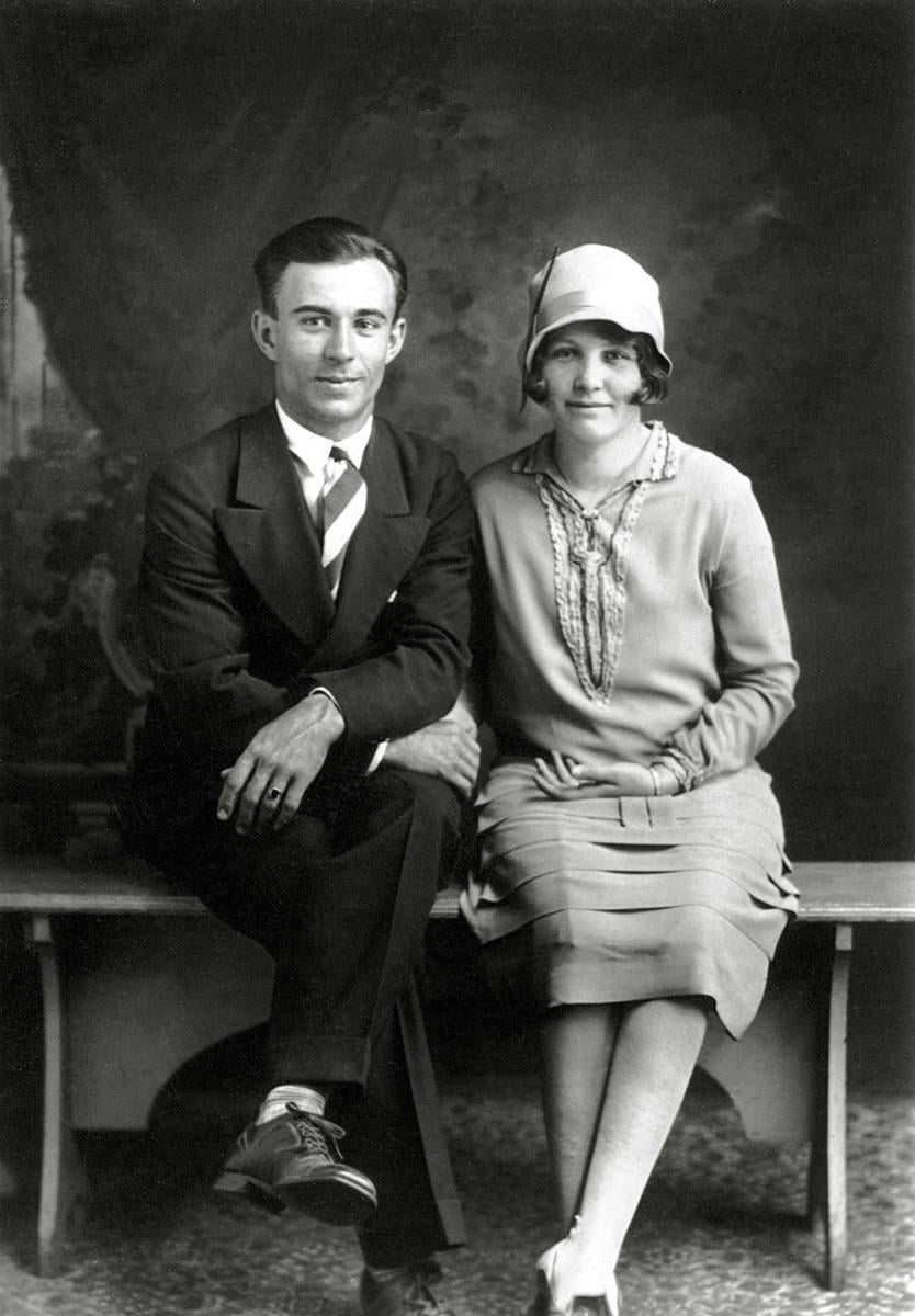 A black and white vintage photo of a man in a suit and tie sitting next to a woman in a cloche hat and 1920s dress, both smiling and posing for a formal portrait on a bench.