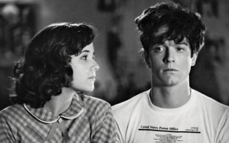 A young woman in a checkered dress with a white collar looks at a young man with messy hair wearing a "U.S. Patent Office" T-shirt. Both appear serious in this black and white image.
