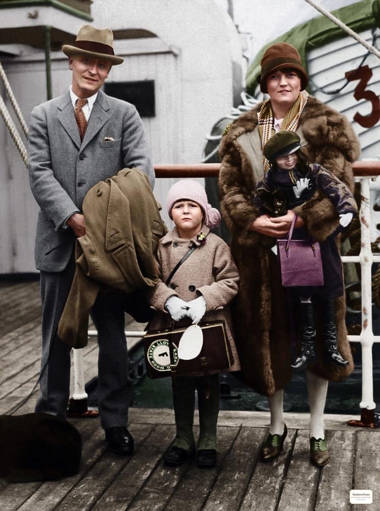 A man in a gray suit, a young girl in a tan coat, and a woman in a brown fur coat with a small dog pose on the deck of a ship, holding luggage and bundled up for travel.